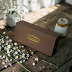 Brown box with 'The Natural B' logo on a wooden surface with flowers and a jar.