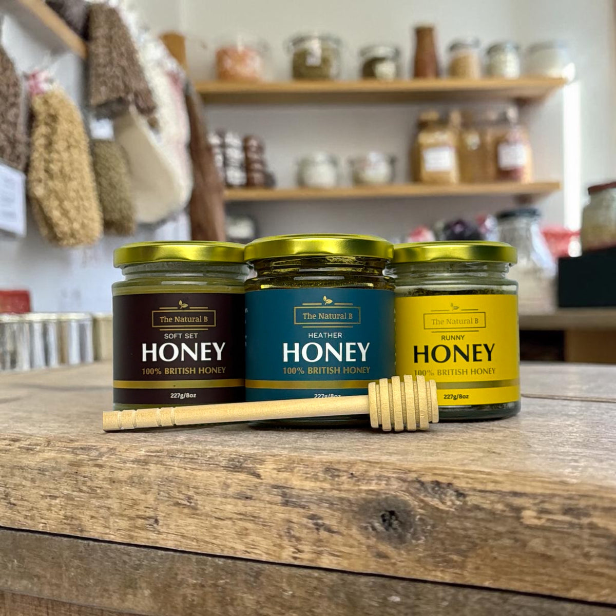 Three jars of honey with labels on a wooden surface, with shelves in the background.