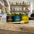 Three jars of honey with labels on a wooden surface, with shelves in the background.