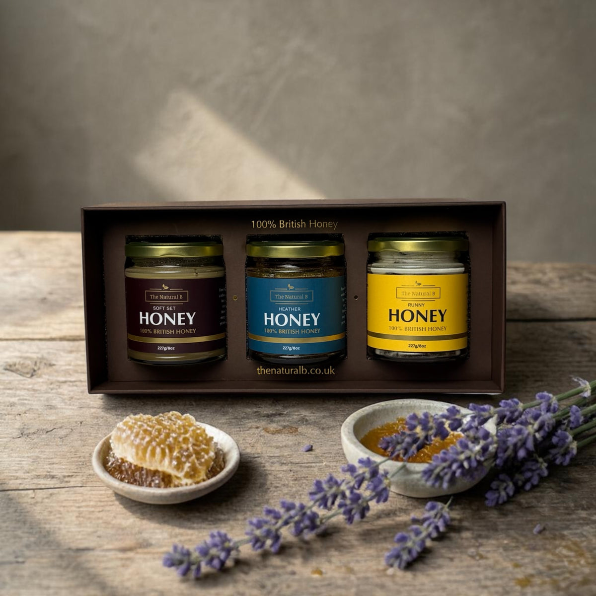 Three jars of honey in a box with honeycomb and lavender on a wooden surface.