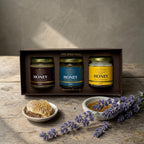 Three jars of honey in a box with honeycomb and lavender on a wooden surface.