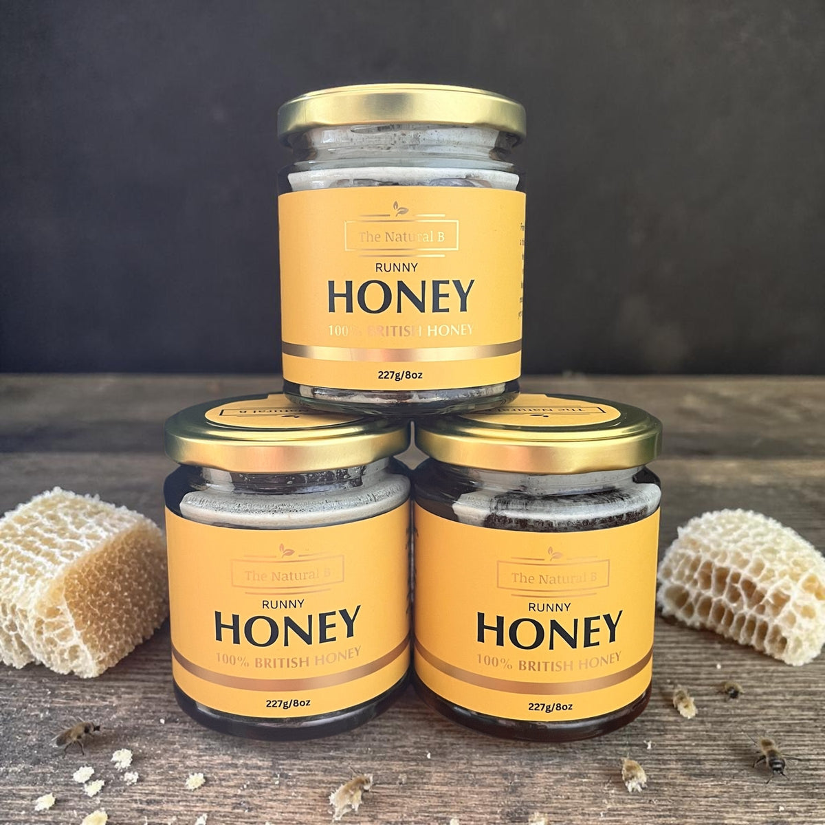 Three jars of honey with gold lids on a wooden surface with honeycomb.