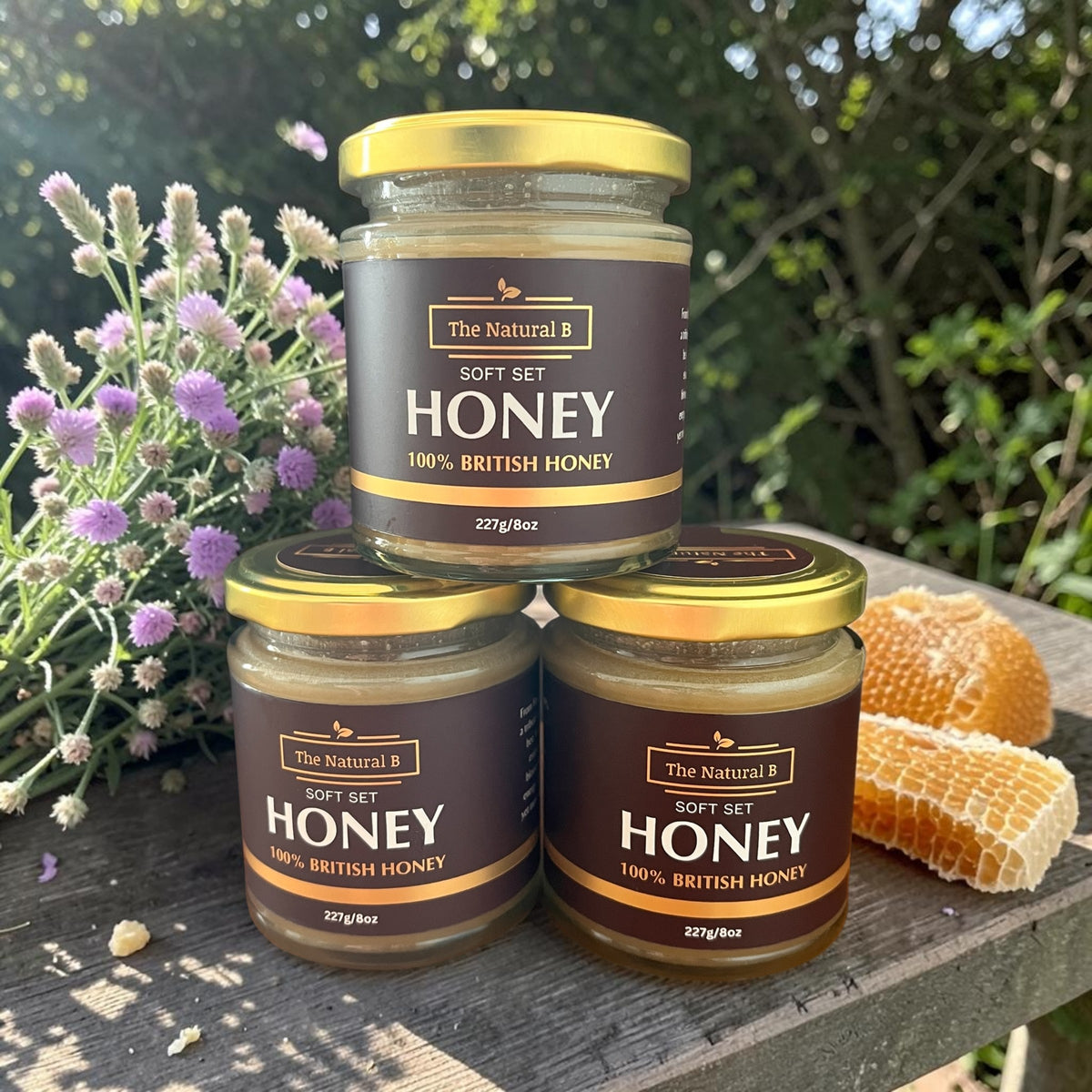 Three jars of 'The Natural B' honey on a wooden surface with flowers and honeycomb in the background.