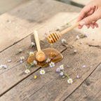 Honey being drizzled from a wooden honey dipper onto a wooden surface with flowers.
